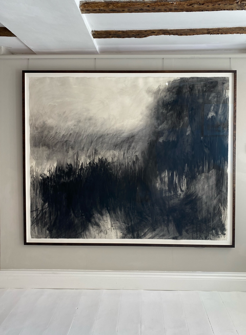 black and white charcoal drawing of abstracted landscape, mounted on white card and framed with thin brown wooden frame. the picture hangs on the wall of a gallery with painted white floor boards, cream walls and white painted ceiling with dark wooden beams. The artwork is moody and atmospheric